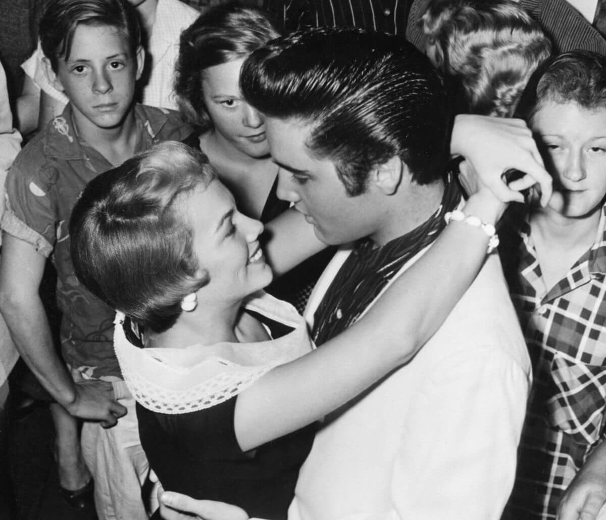 A black and white picture of Elvis and Anita Wood dancing in a crowd of people. She has her arms around his neck.