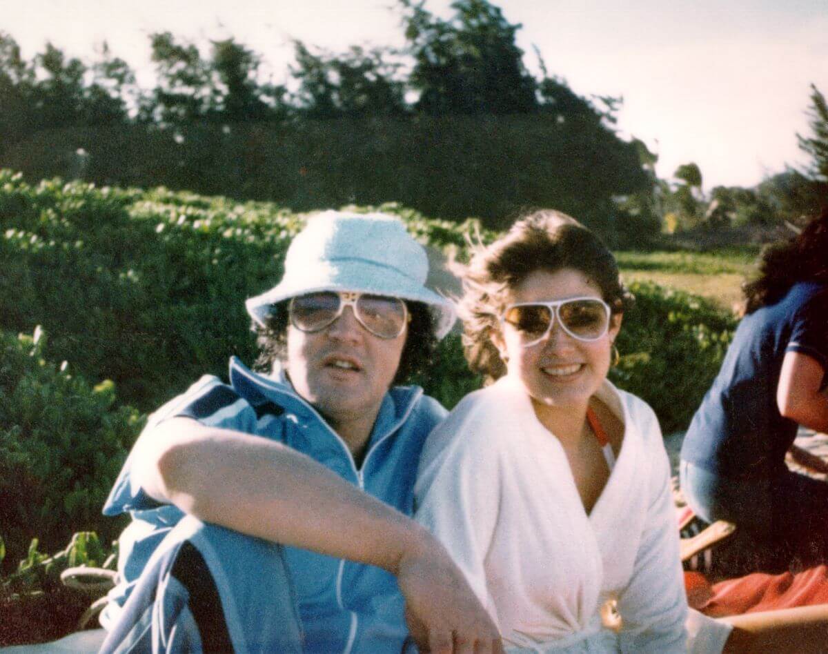Elvis wears a bucket hat and sunglasses and sits next to Ginger Alden, who wears sunglasses. They are outside.