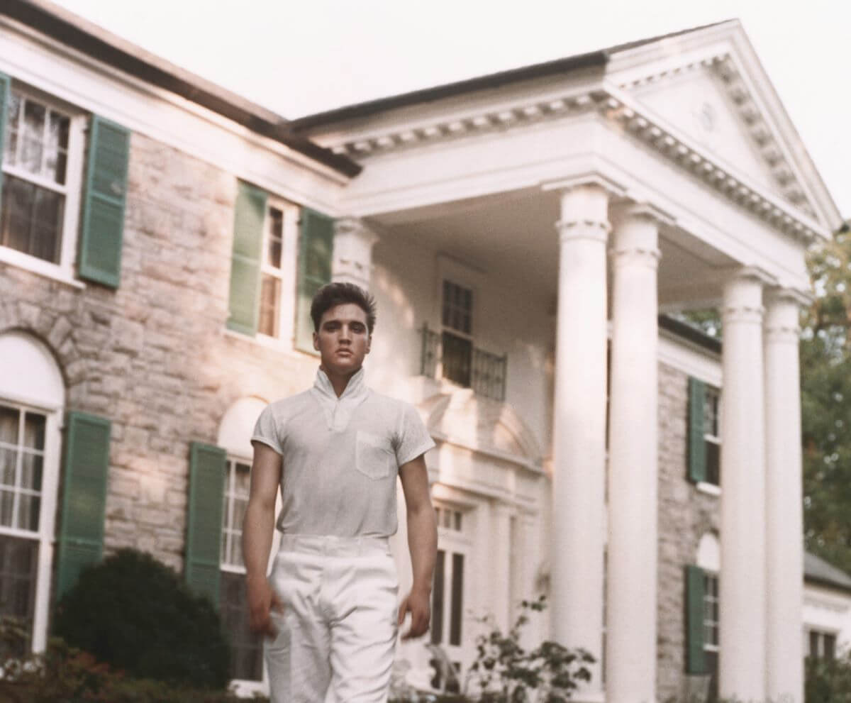 Elvis Presley wears a white shirt and white pants. He walks in front of Graceland.