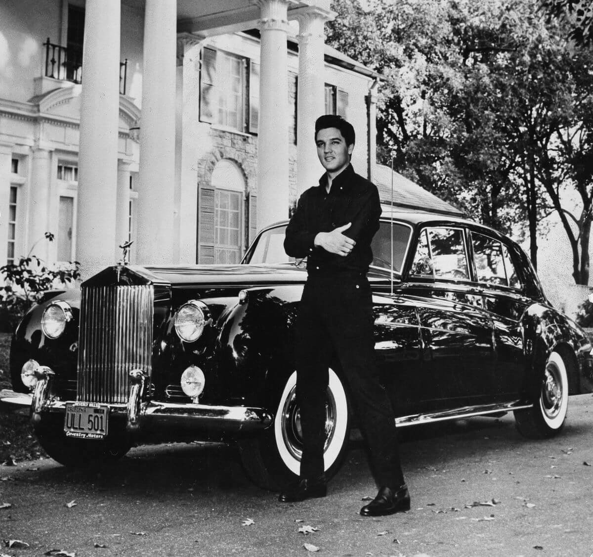 A black and white picture of Elvis standing with his arms crossed. There is a car behind him that is parked in front of Graceland.