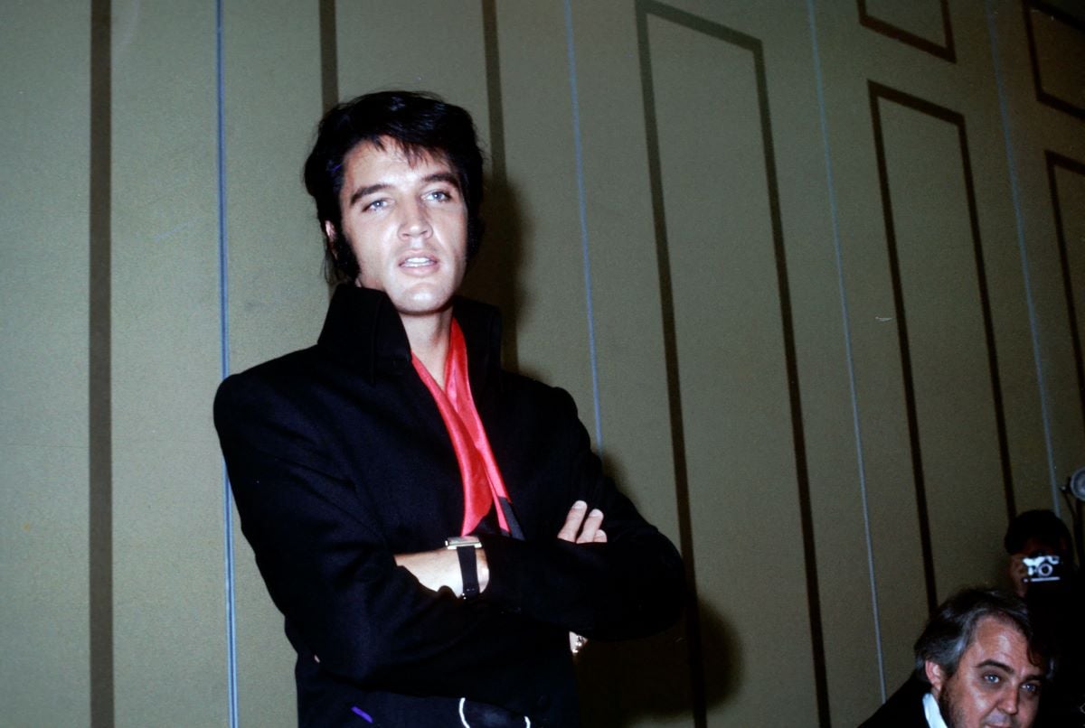Elvis wears a black jacket with a red tie and stands with his arms crossed.