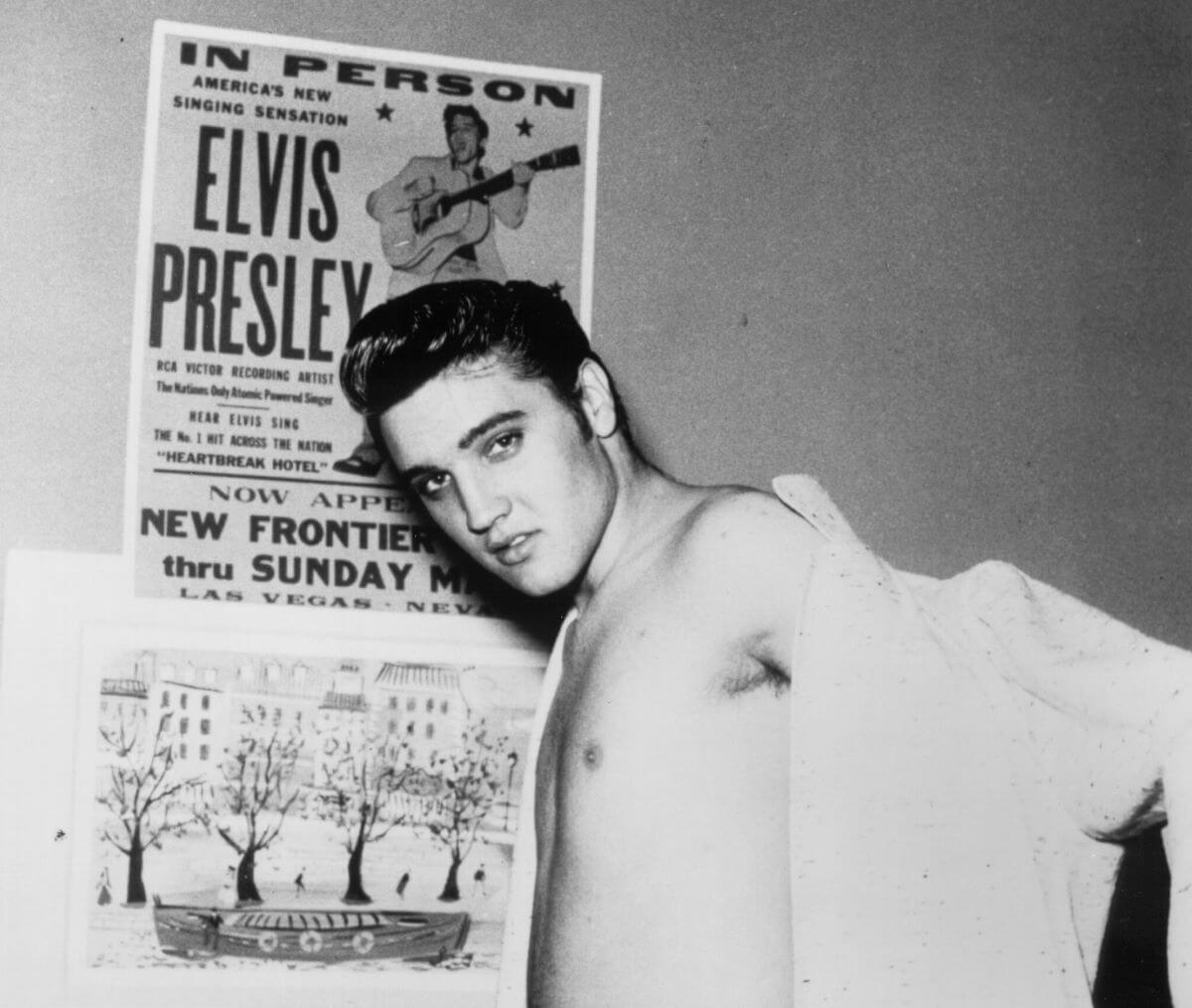 A black and white picture of Elvis putting his shirt on and standing in front of a poster for his show at the New Frontier Hotel.