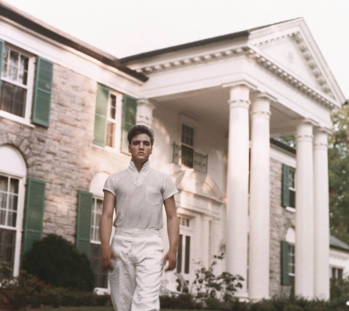 Elvis wears a white shirt and white pants. He stands in front of Graceland.
