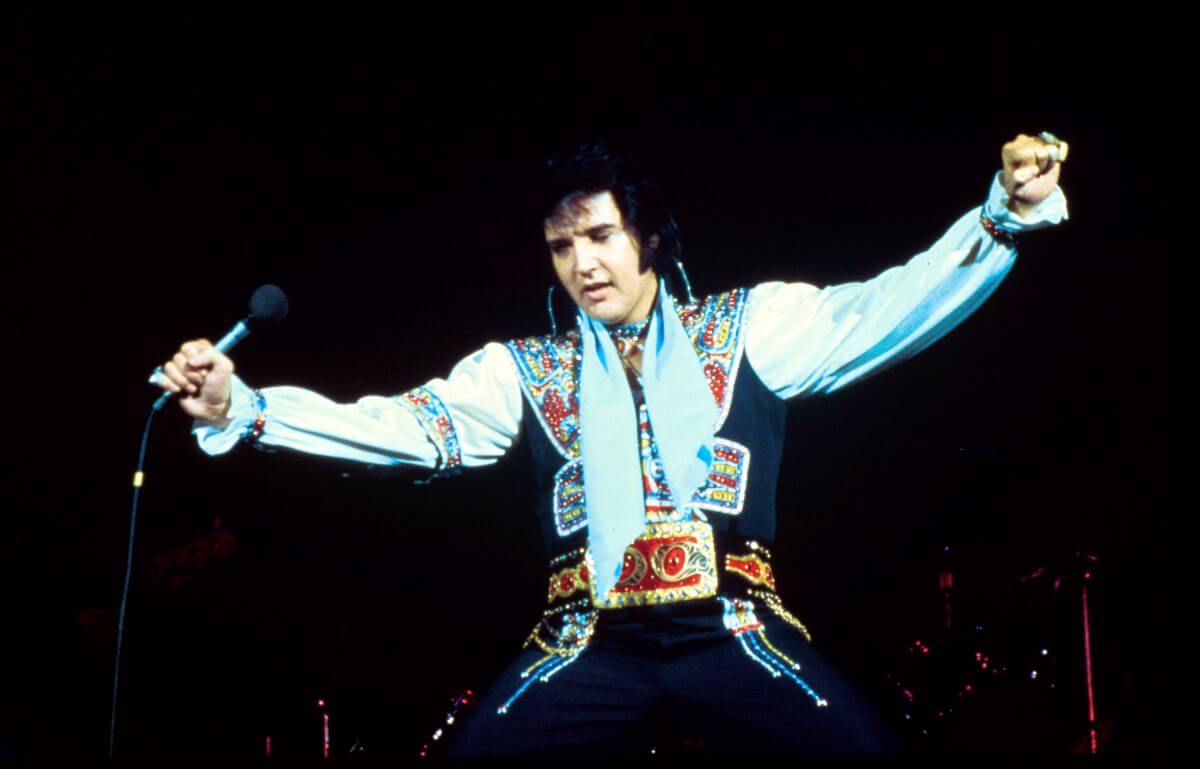 Elvis Presley wears a sparkly vest and belt during a performance. He holds his arms wide.