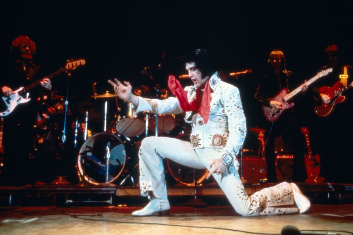 Elvis Presley lunges onstage while wearing a white jumpsuit and red scarf.