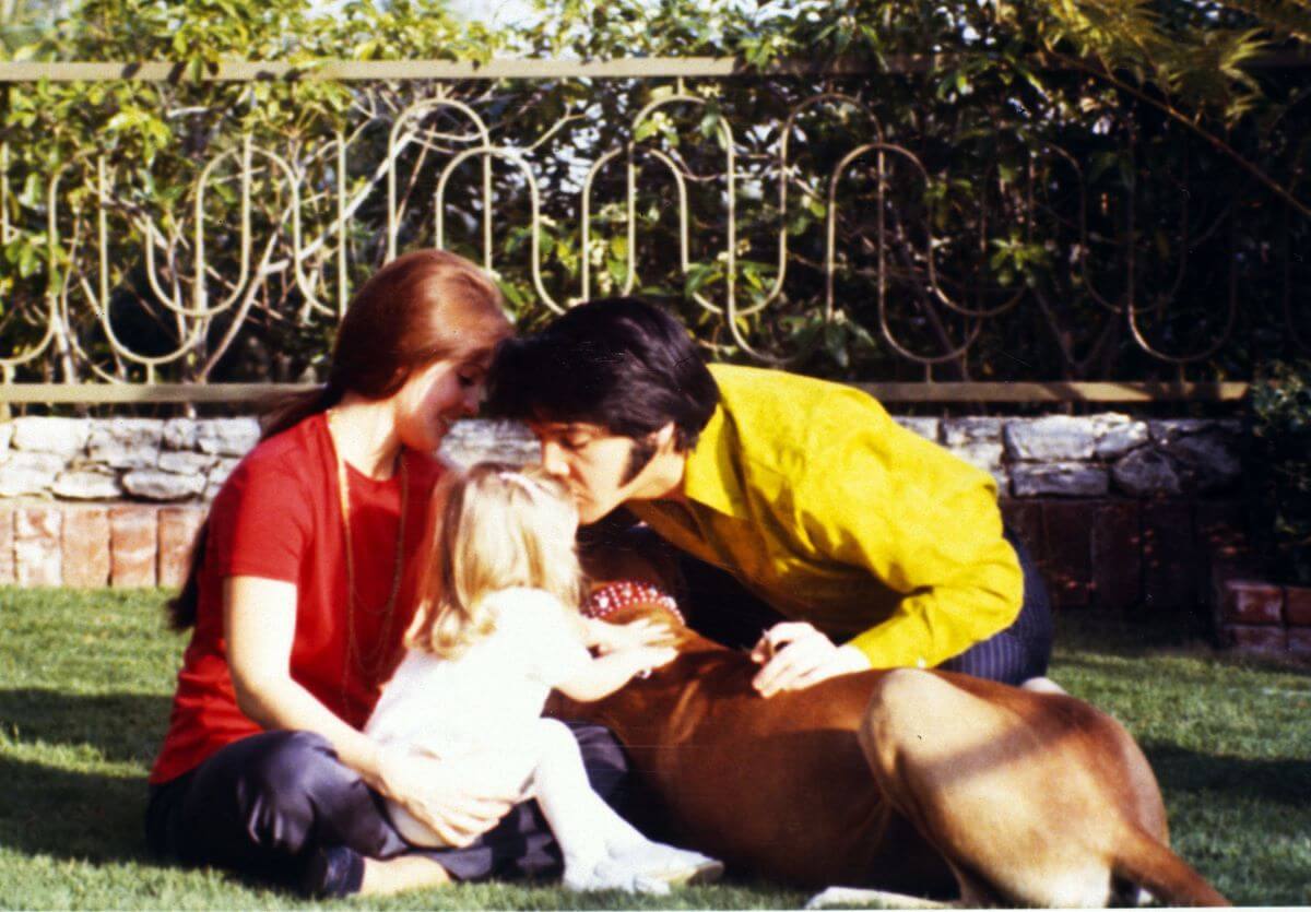 Priscilla, Lisa Marie, and Elvis Presley sit on a lawn surrounding a dog.
