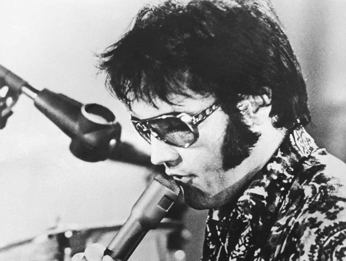 A black and white picture of Elvis wearing sunglasses and singing into a microphone.