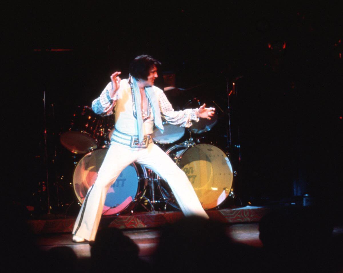 Elvis wears a white jumpsuit and stands onstage.