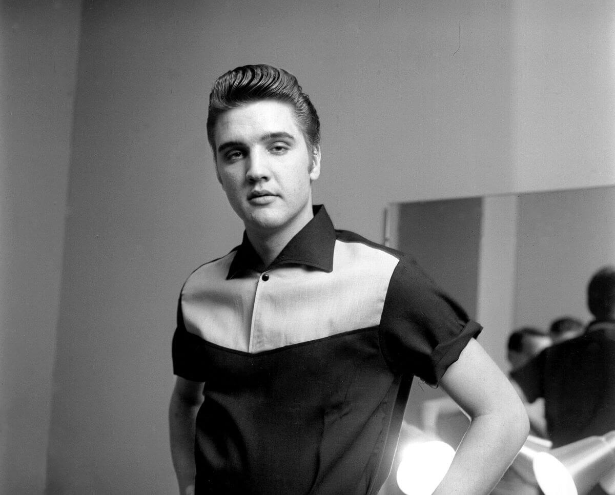 A black and white picture of Elvis Presley standing in a polo shirt with his hands on his hips.