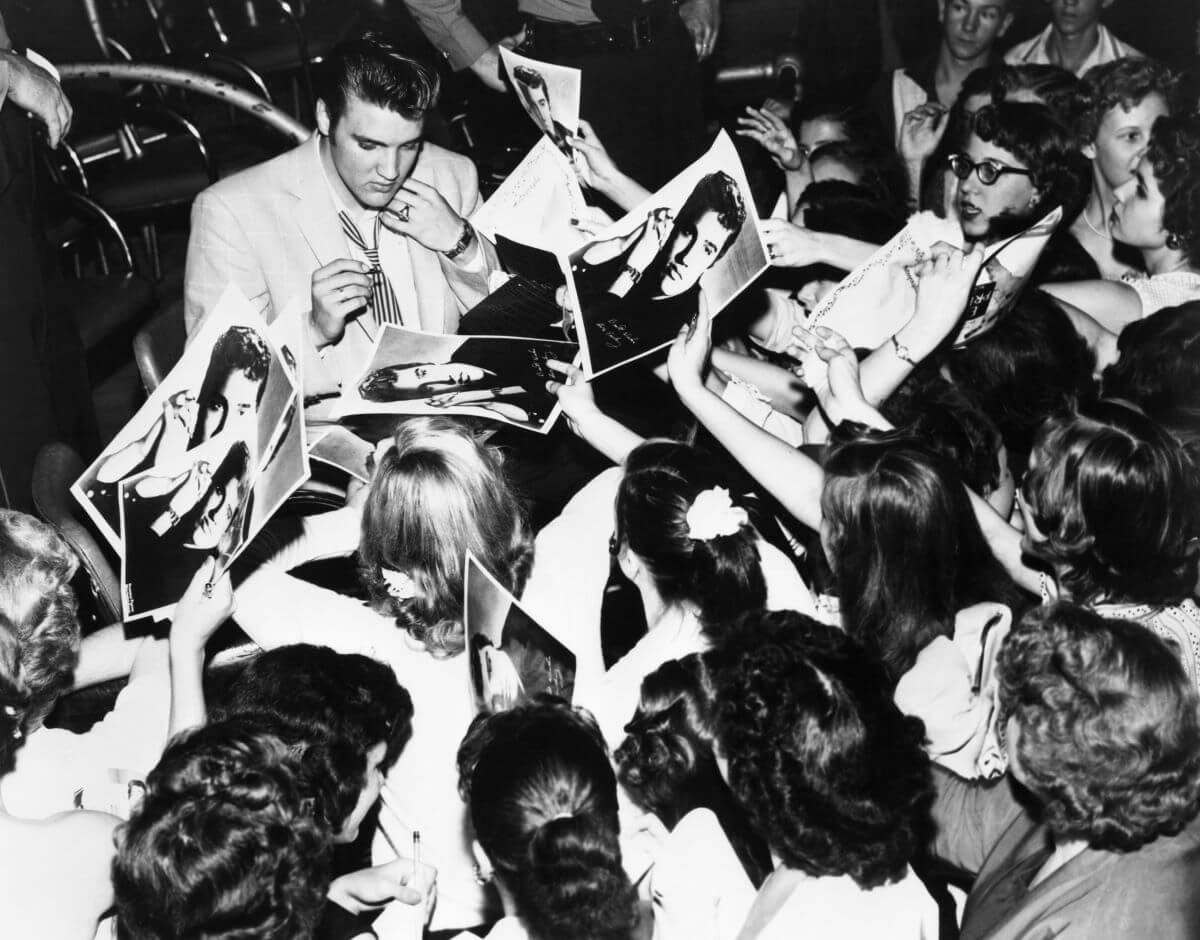 A black and white picture of Elvis signing pictures of himself for a group of fans.