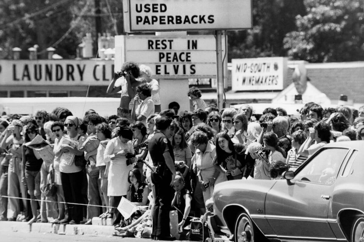 A black and white picture of crowds fathered under a sign that says Rest in Peace Elvis. They stand near a dry cleaner's and a wine store. There is a car in the bottom right of the image.