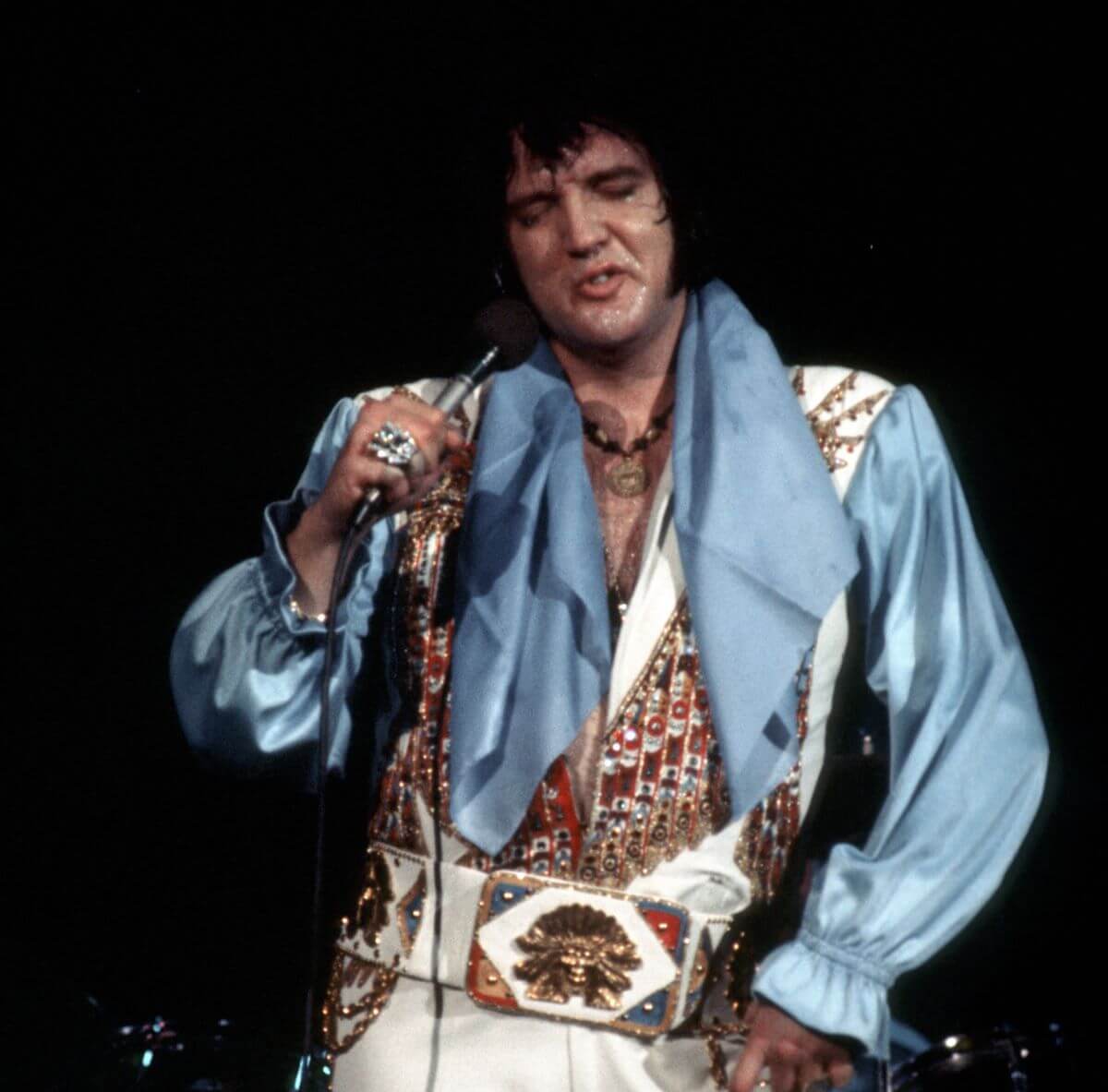 Elvis Presley wears a blue shirt and a bejeweled vest and belt. He sings into a microphone.