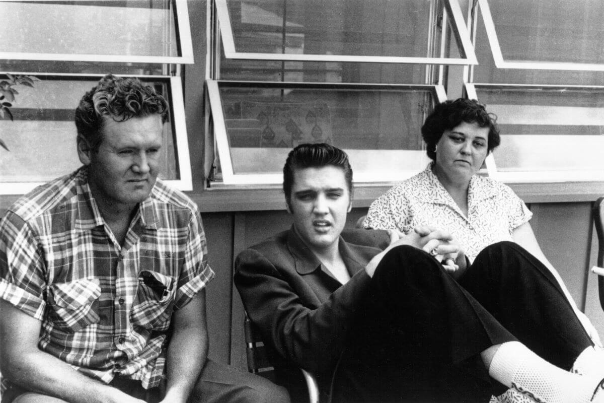 A black and white picture of Elvis and his parents sitting outside a building with opened windows. Elvis leans back and lifts his knees towards his body.