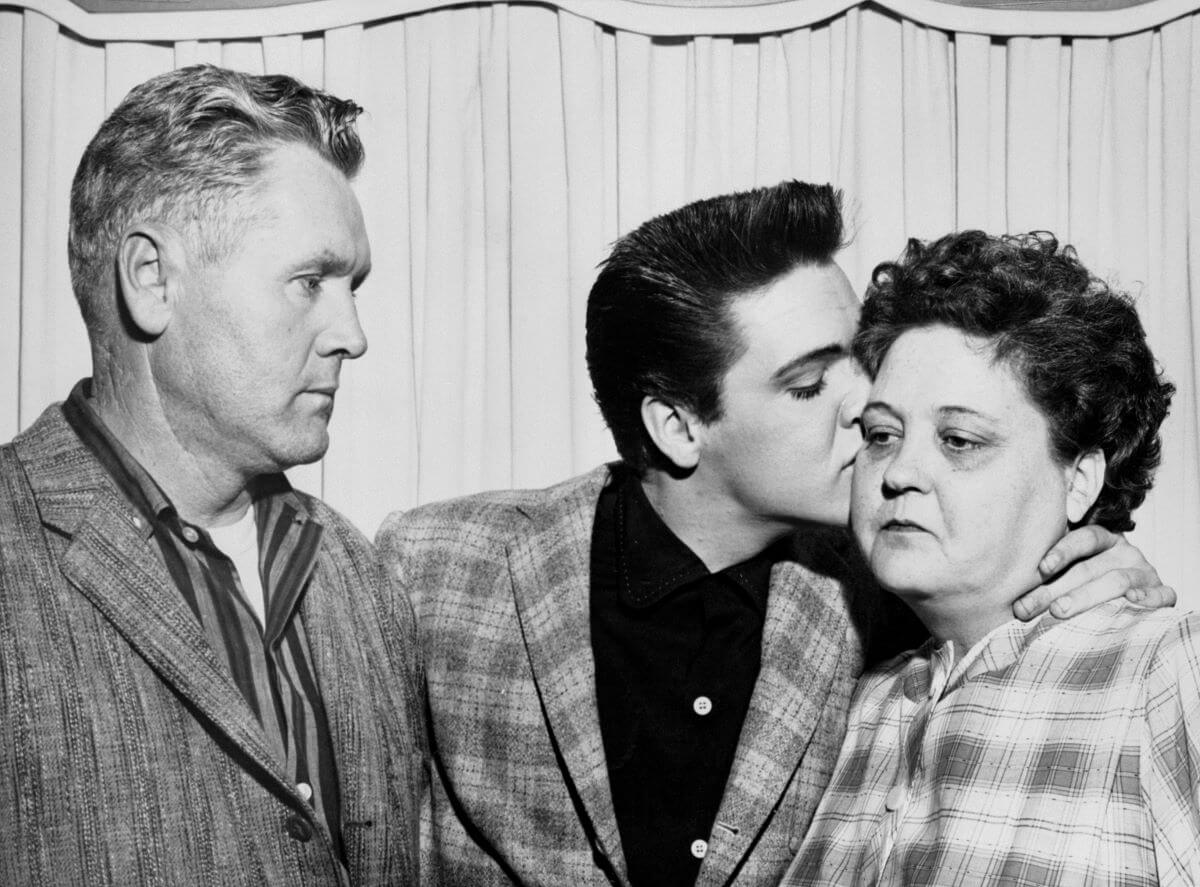 A black and white picture of Elvis Presley kissing his mother's cheek. His father watches on.