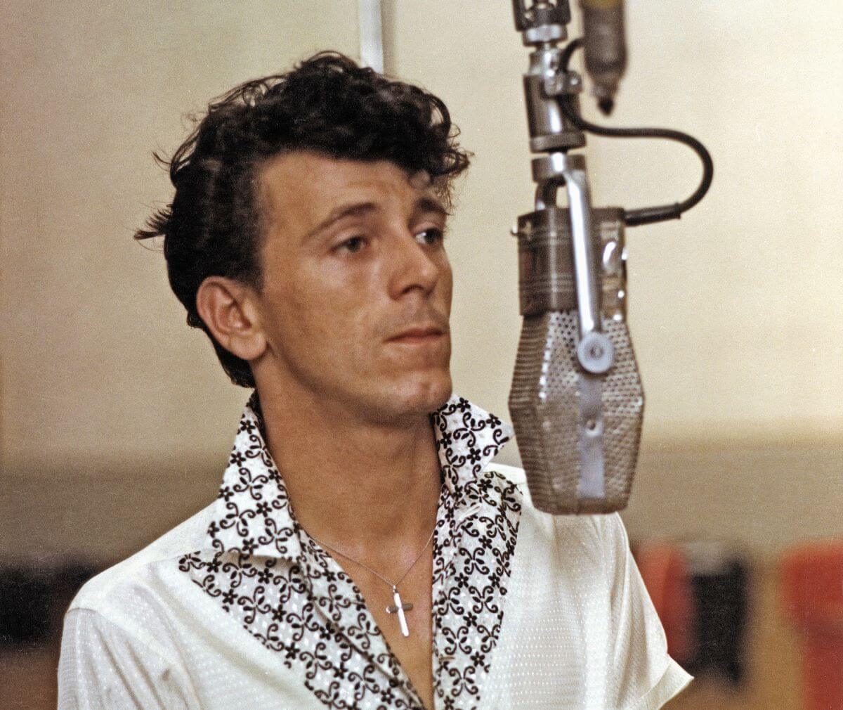Gene Vincent wears a white shirt with a black and white patterned collar and a silver cross necklace. He stands in front of a microphone.