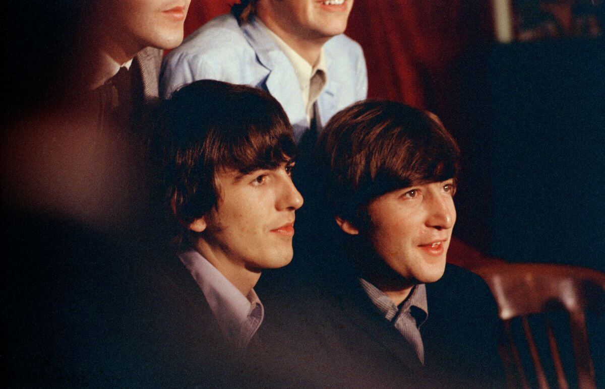 George Harrison and John Lennon wear suits and sit together. Paul McCartney and Ringo Starr are behind them.