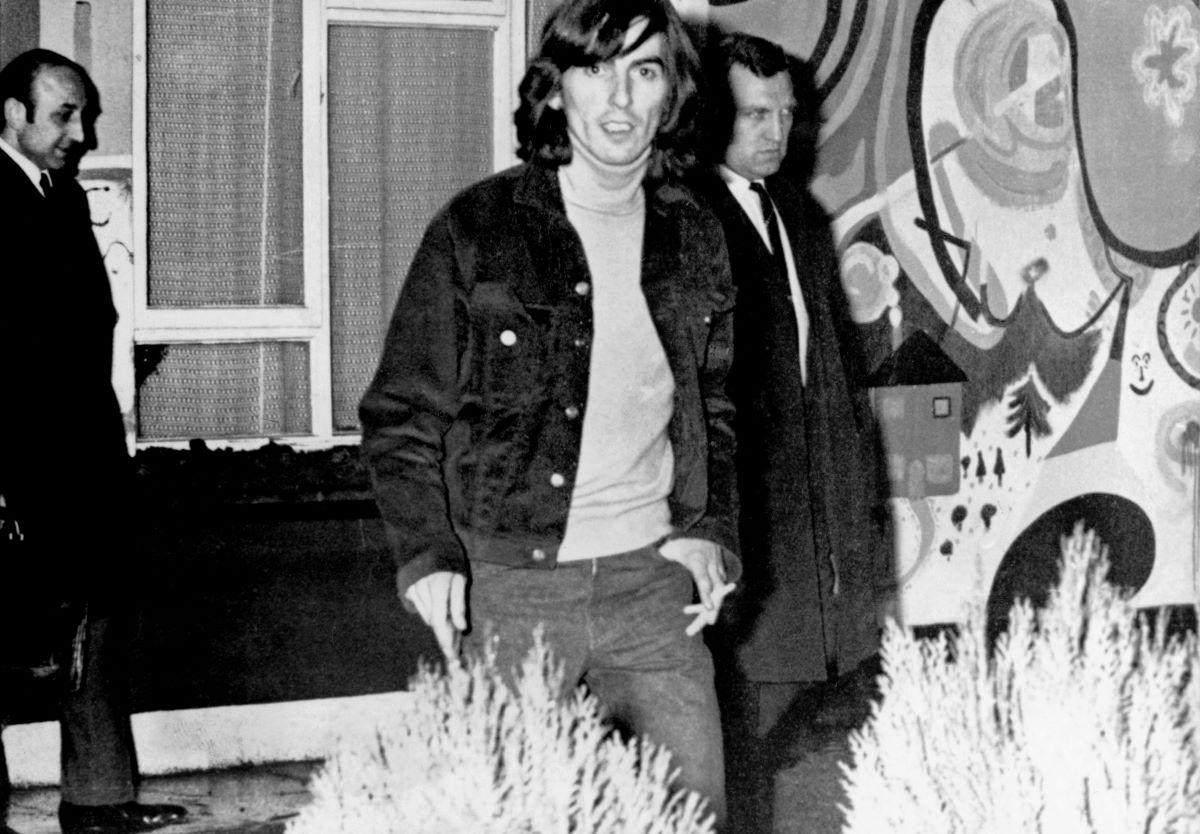 A black and white picture of George Harrison walking out of his house while police officers stand behind him.