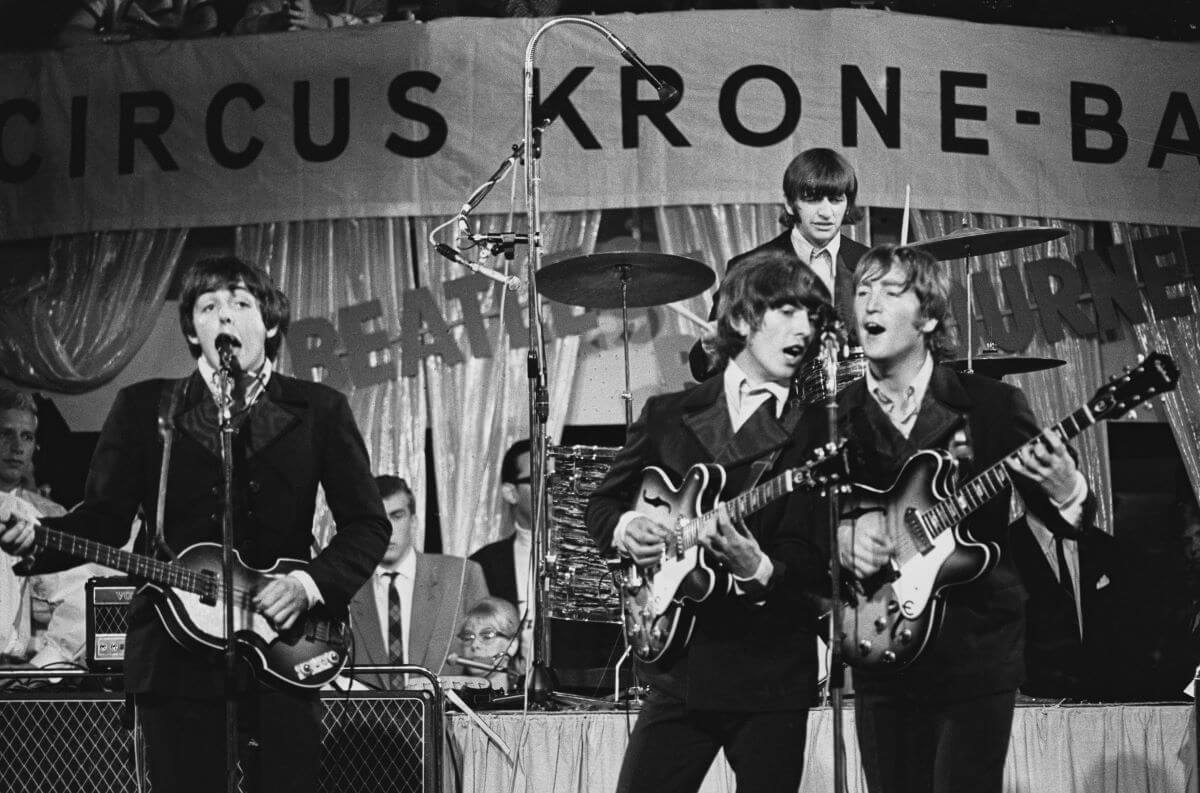 A black and white picture of The Beatles during a concert. Paul McCartney plays guitar and sings, George Harrison and John Lennon play guitars and sing into the same microphone. Ringo Starr drums on a platform behind them.