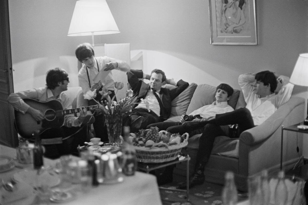 John Lennon, George Harrison, Brian Epstein, Ringo Starr, and Paul McCartney sit on a couch. Lennon holds a guitar.