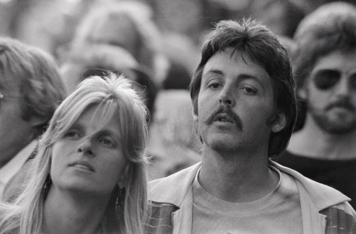 A black and white picture of Linda and Paul McCartney standing in the crowd at a concert. McCartney has a mustache.