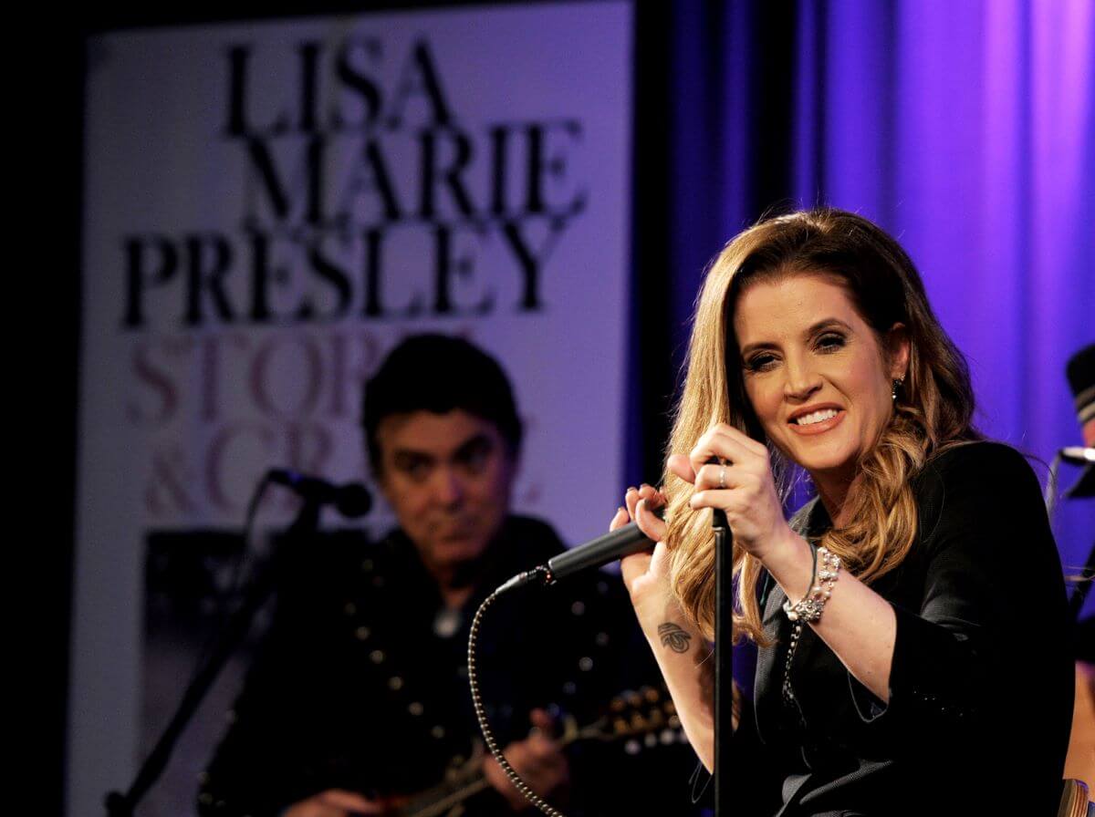 Lisa Marie Presley holds a microphone and smiles. She sits in front of a sign with her name on it lit in purple light.