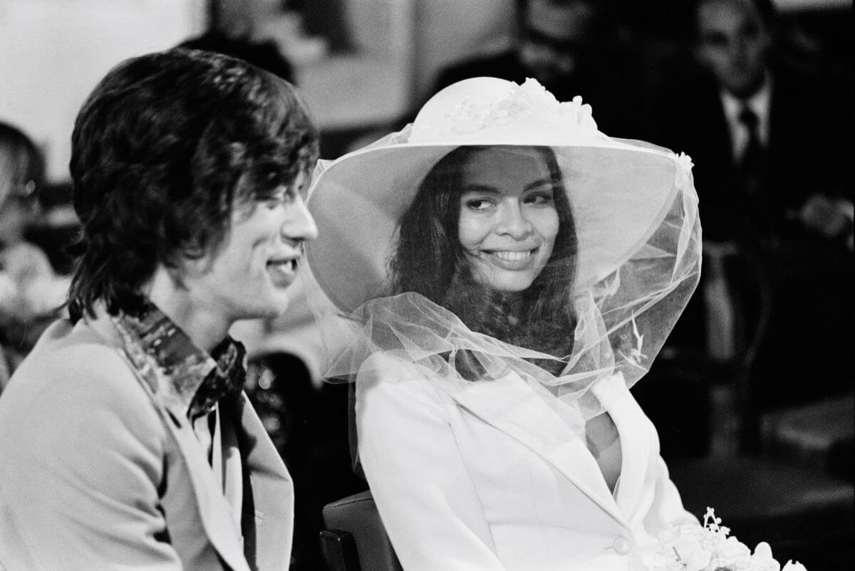 A black and white picture of Mick Jagger sitting next to Bianca Jagger, who wears a hat and a veil.