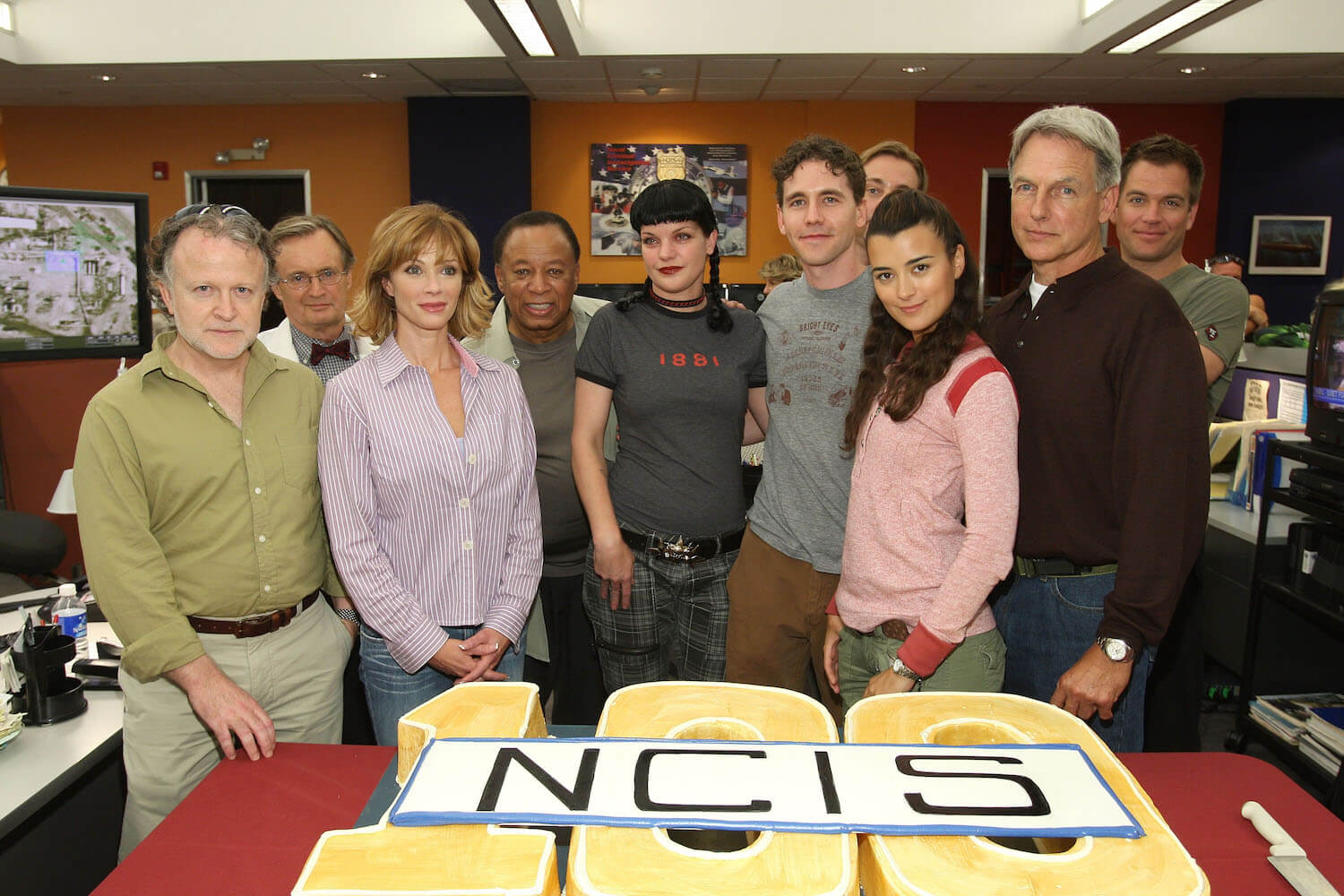 'NCIS' cast members with producers Shane Brennan (L) and Charles Floyd Johnson standing in front of a cake celebrating the 100th episode