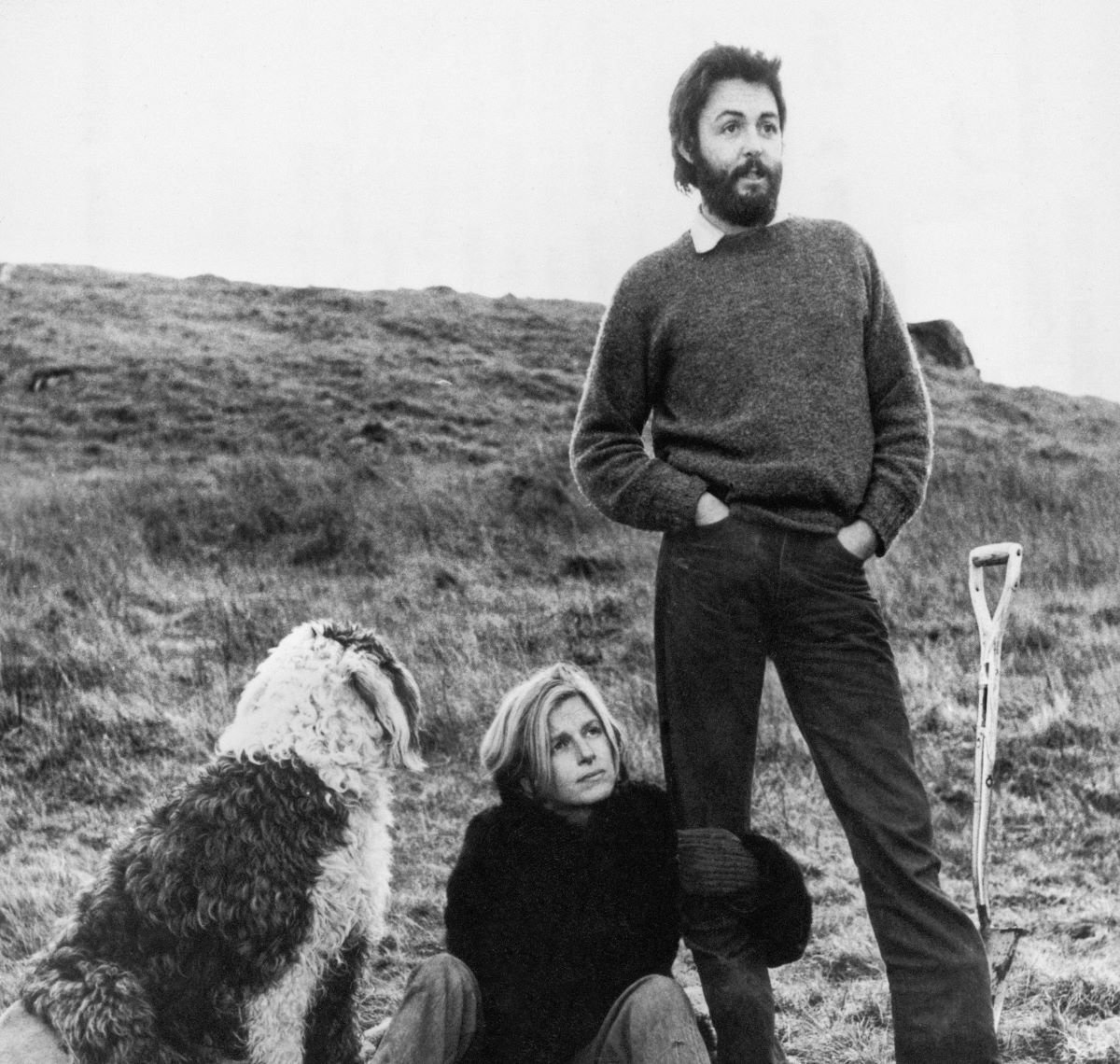 A black and white picture of Paul McCartney standing next to a shovel. Linda McCartney sits on the ground between Paul and a dog.