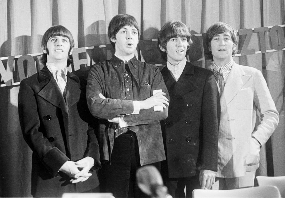 A black and white picture of Ringo Starr, Paul McCartney, George Harrison and John Lennon of The Beatles standing in a line in front of a curtain. McCartney has his arms crossed.