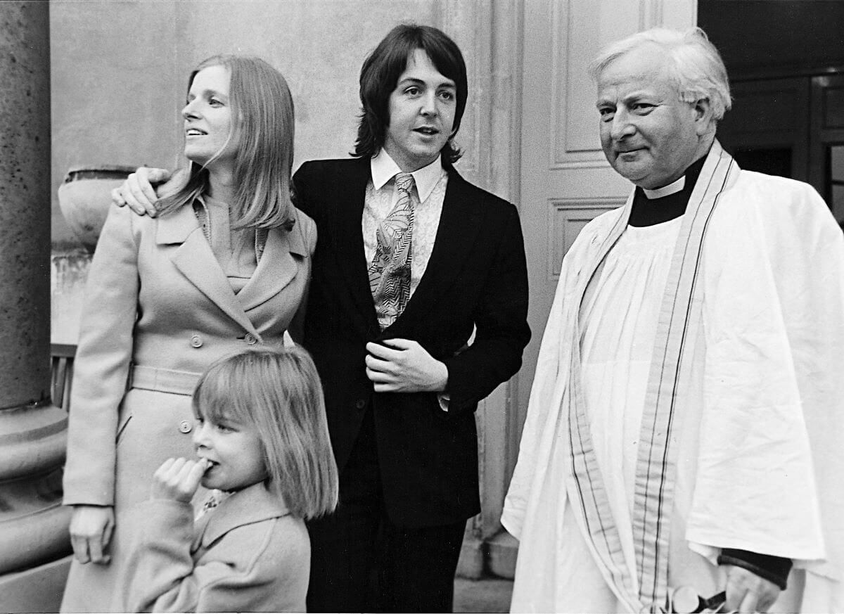 Paul McCartney stands with his arm around Linda McCartney's shoulders on their wedding day. Their daughter Heather stands in front of them and a priest stands to the side.