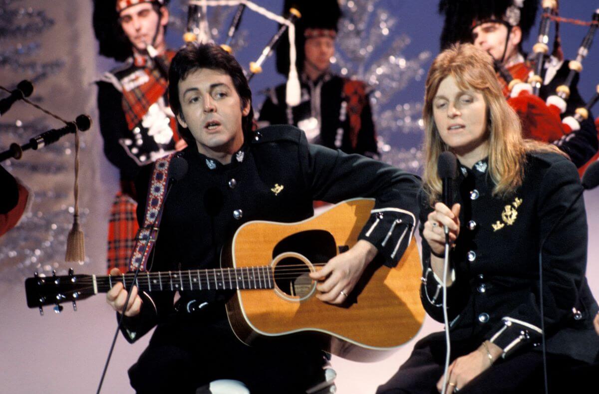 Paul McCartney plays guitar and sings with Linda McCartney, who holds a microphone. Three bagpipers stand behind them amongst silver Christmas trees.
