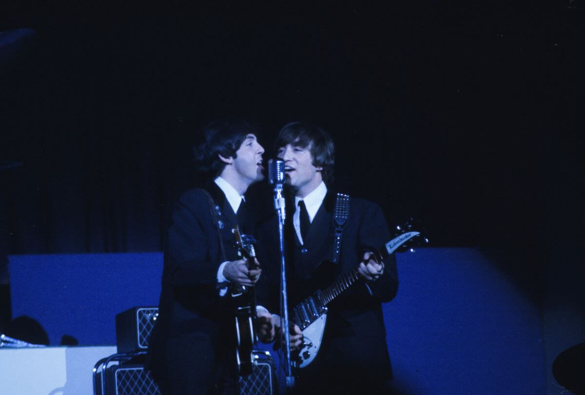 Paul McCartney and John Lennon play guitars and sing into the same microphone.