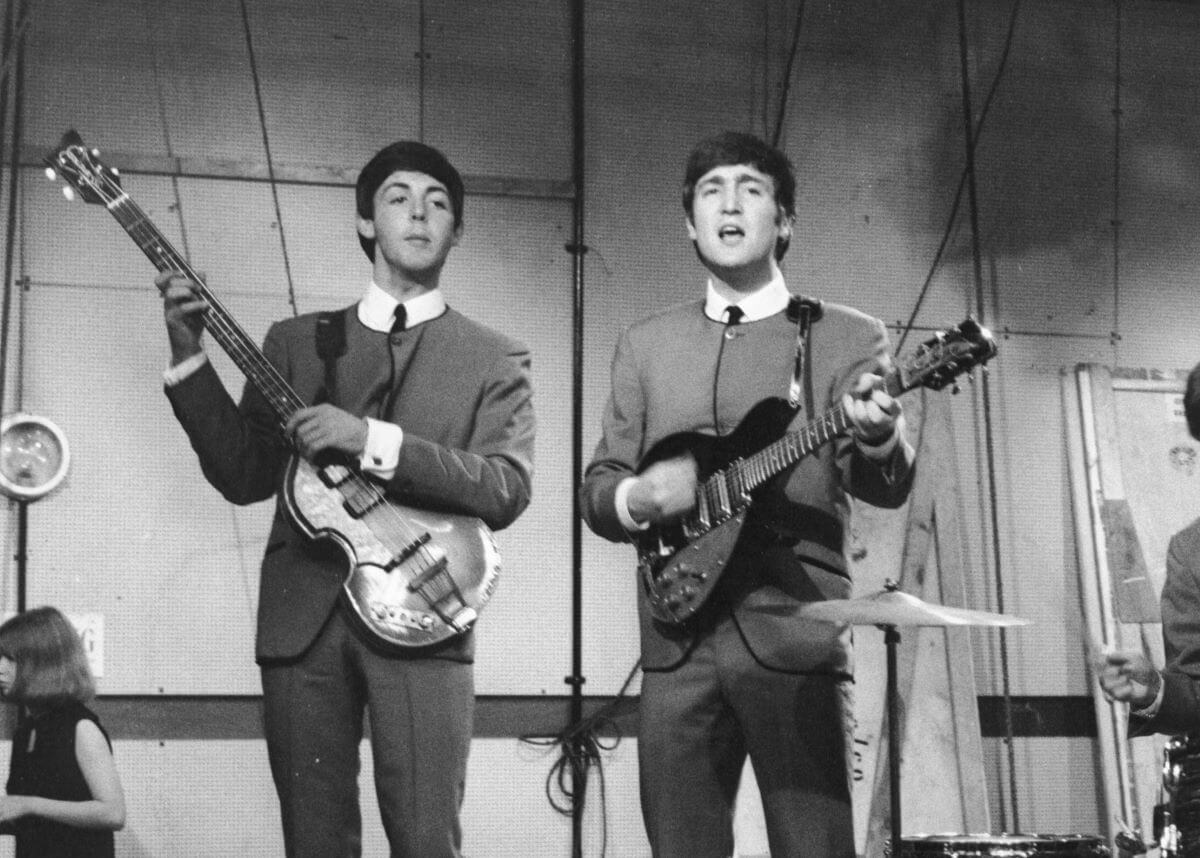A black and white picture of Paul McCartney and John Lennon wearing matching suits and playing guitars. They sing.