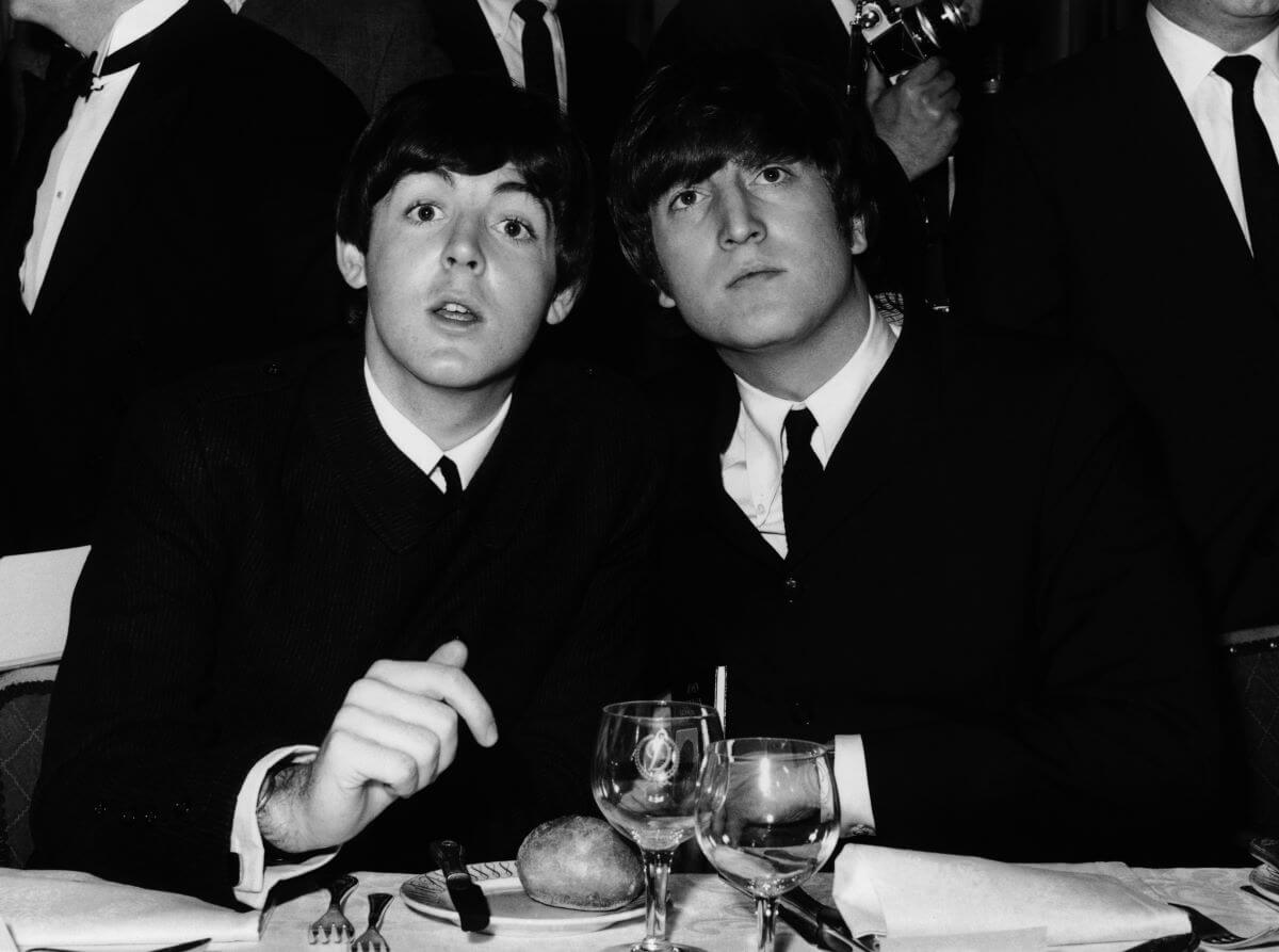 A black and white picture of Paul McCartney and John Lennon wearing suits and sitting at a dinner table.