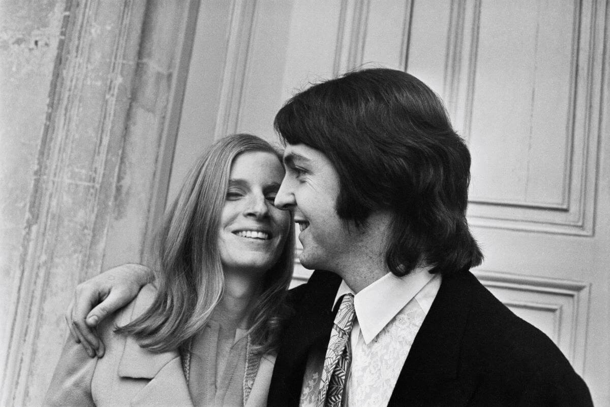 A black and white picture of Linda and Paul McCartney. He has her arm around his shoulders and they both smile.
