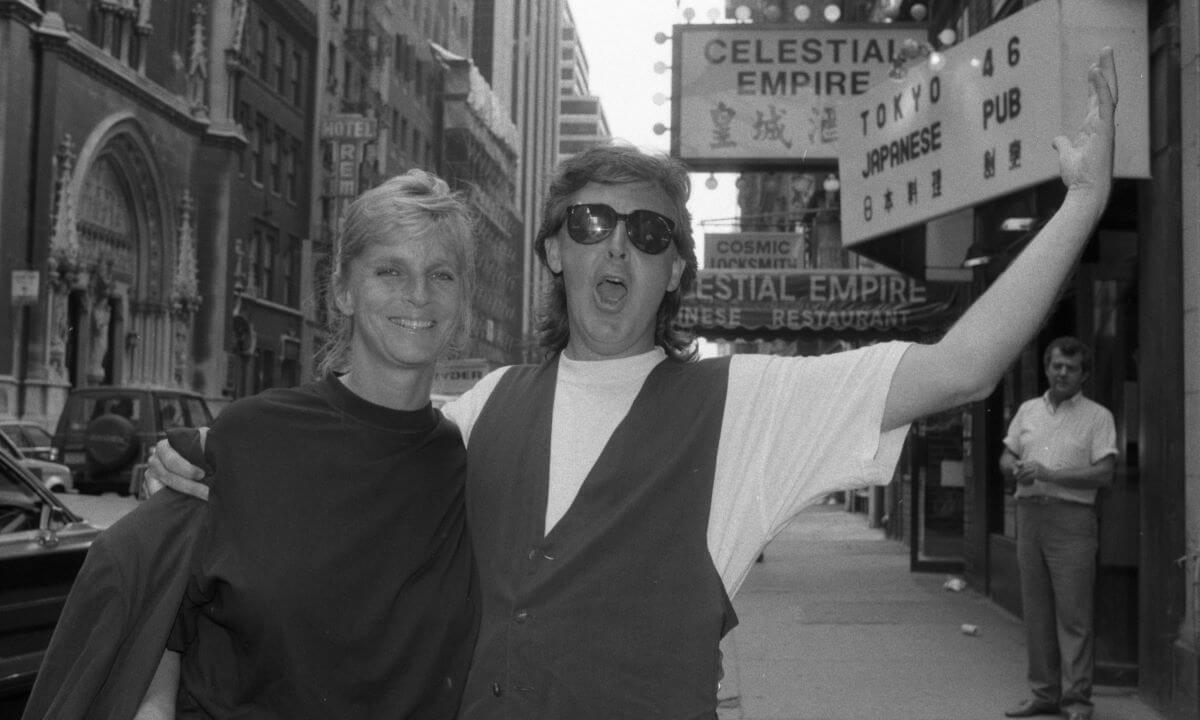 A black and white picture of Paul and Linda McCartney. He has his arm around her shoulders and holds the other up in the air. He wears sunglasses.