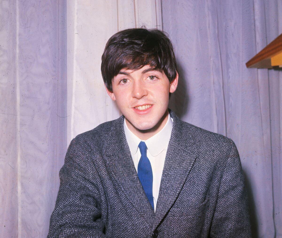 Paul McCartney wears a blue suit and sits in front of a curtain.