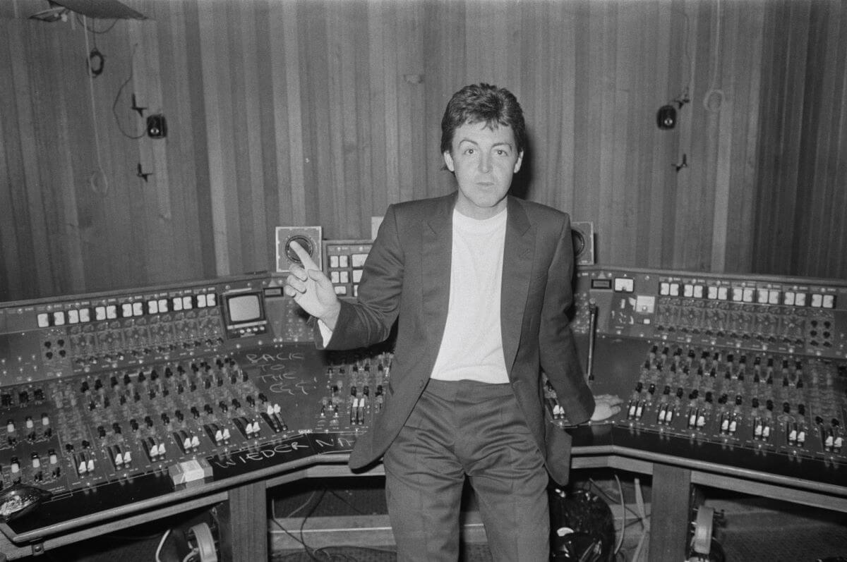 A black and white picture of Paul McCartney holding up a hand as he leans against a control panel.