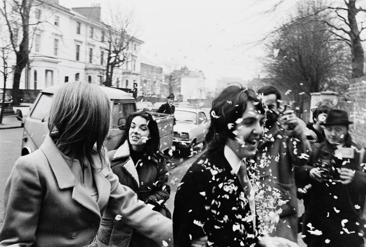 A black and white picture of Paul and Linda McCartney on their wedding day. She holds his arm and he walks into confetti.