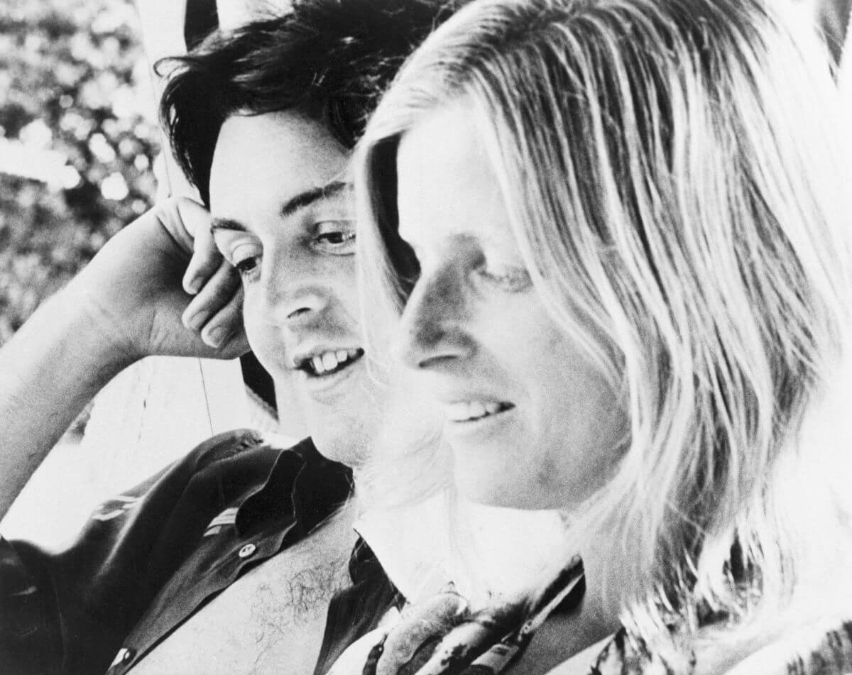 A black and white picture of Paul and Linda McCartney sitting together. He leans his head onto his fist and they both smile.