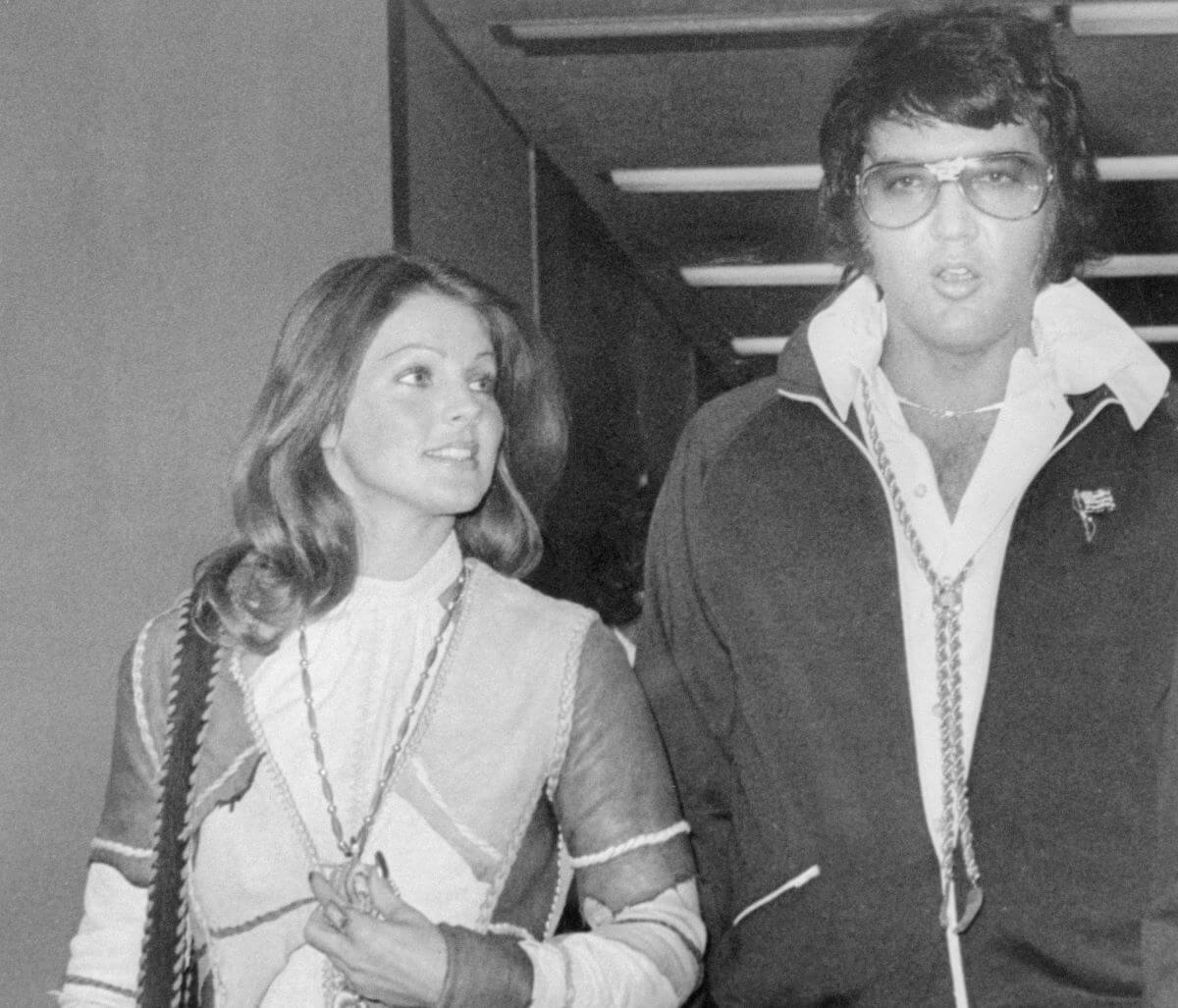 A black and white picture of Priscilla and Elvis Presley walking out of court. She looks towards him and he wears sunglasses.