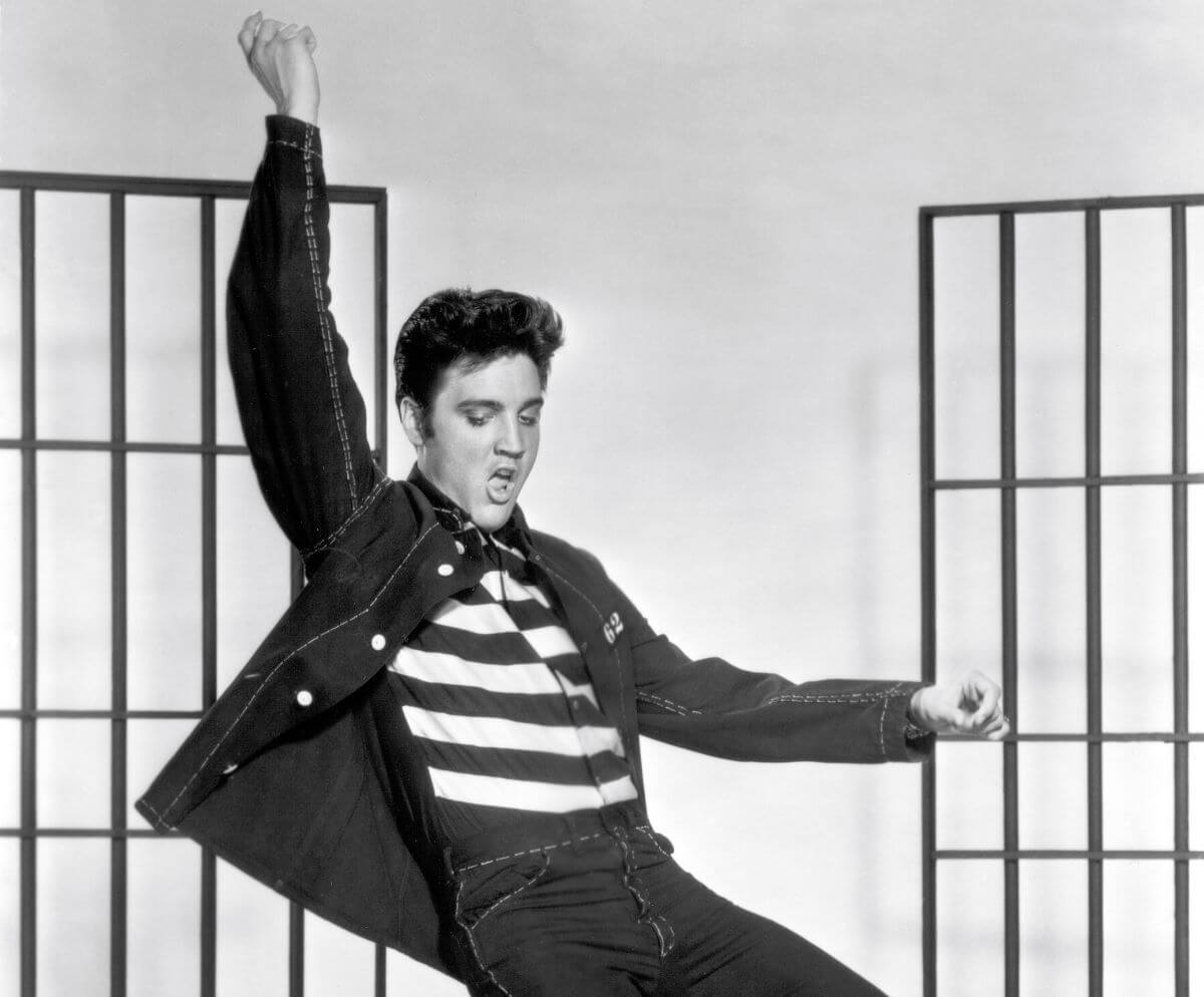 A black and white picture of Elvis wearing a striped shirt. He holds one arm above his head and one out in front of him.