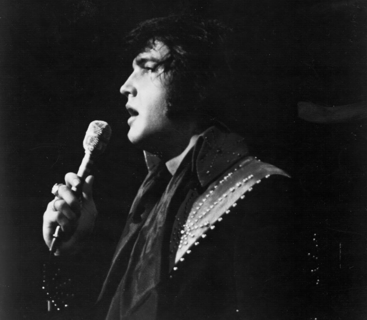 A black and white picture of Elvis in side profile, holding a microphone up to his face.