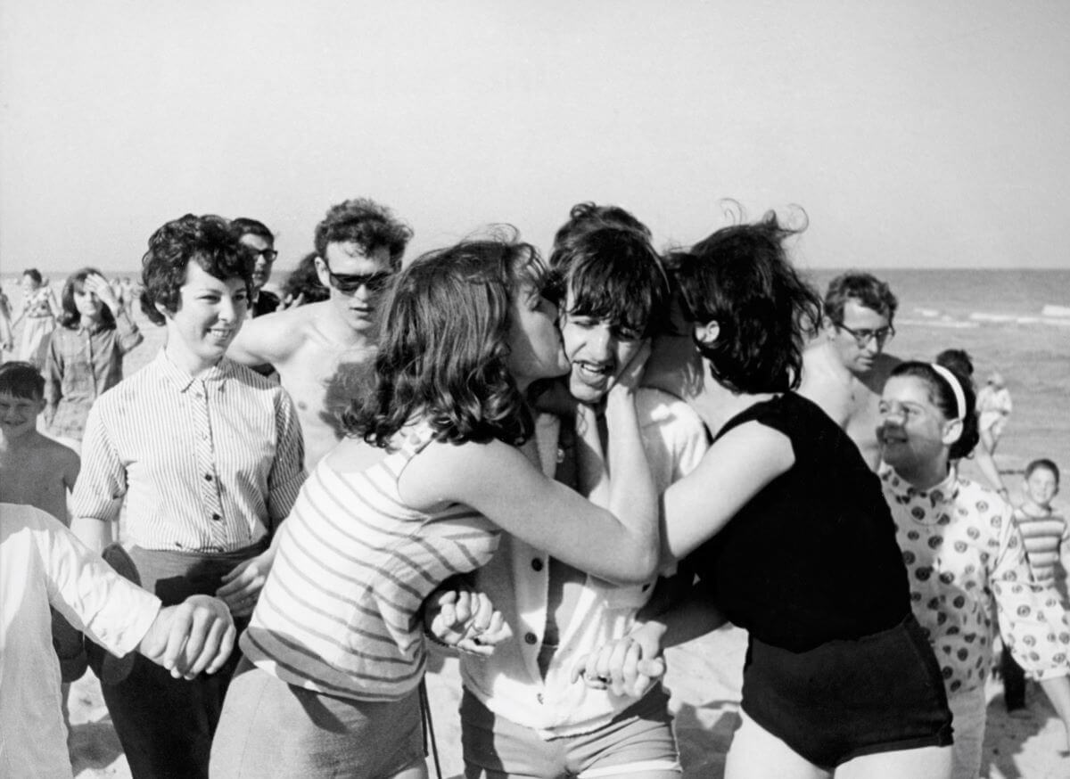 A black and white picture of fans kissing Ringo Starr on the cheeks while on a beach.