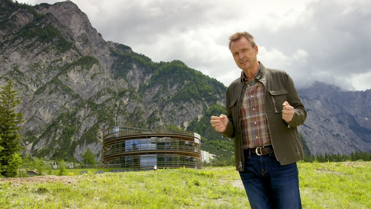 'The Amazing Race' host Phil Keoghan standing in front of a modern building and mountains
