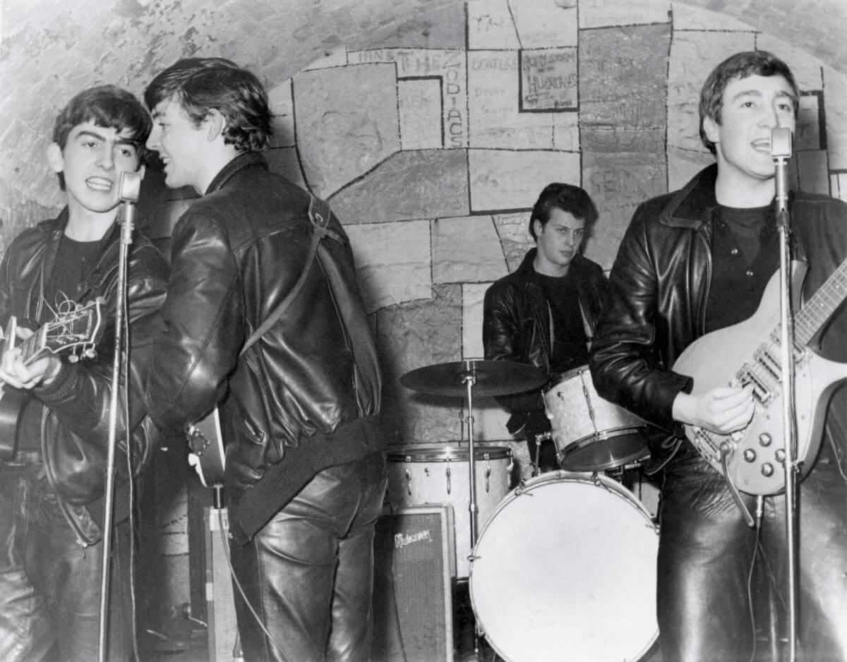 A black and white picture of George Harrison, Paul McCartney, Pete Best, and John Lennon of The Beatles playing a concert in front of a stone wall.
