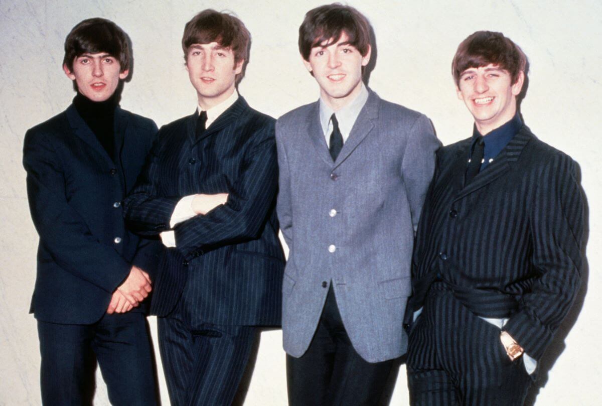 George Harrison, John Lennon, Paul McCartney, and Ringo Starr lean against a white wall. McCartney and Starr smile.