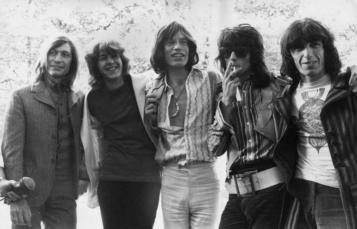 A black and white picture of Charlie Watts, Mick Taylor, Mick Jagger, Keith Richards and Bill Wyman standing with their arms around each other.