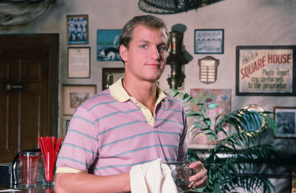 Woody Harrelson posing as Woody Boyd on the set of 'Cheers' wearing a pink shirt while holding a glass and towel.