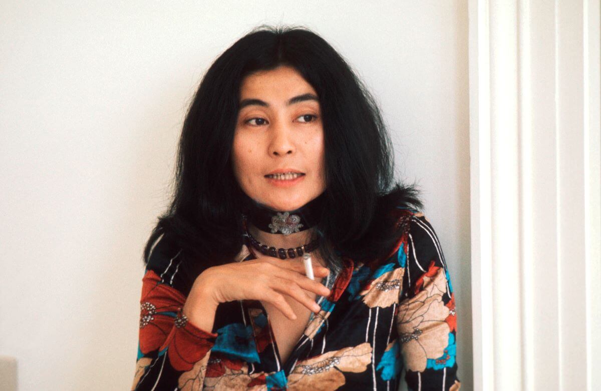 Yoko Ono sits against a white wall and holds a cigarette in her hand.